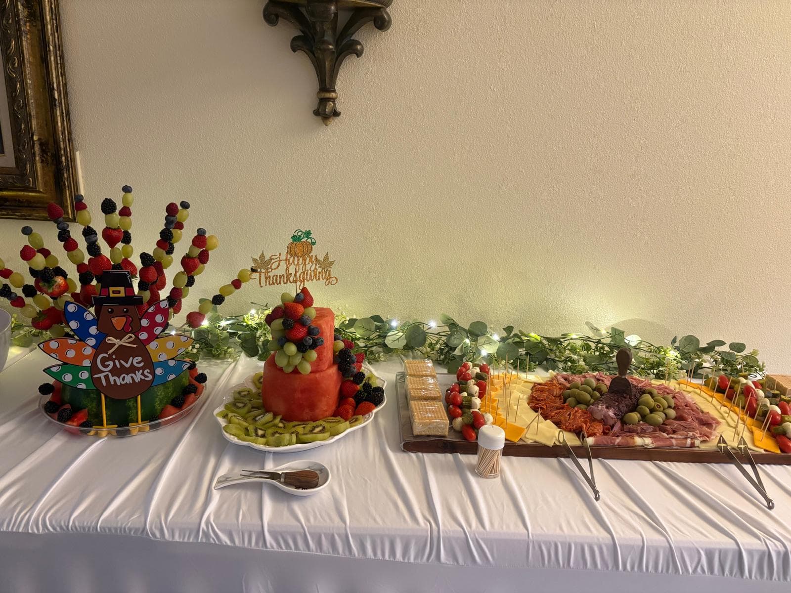 Wide view of a Thanksgiving catering spread with a turkey fruit skewer display, tiered watermelon cake and charcuterie board