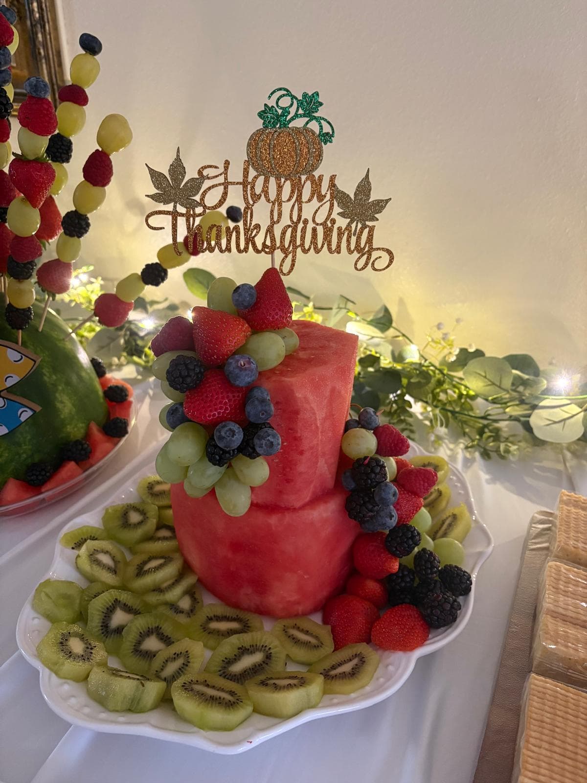 Tiered watermelon fruit cake topped with berries, grapes and a Happy Thanksgiving topper, surrounded by sliced kiwi